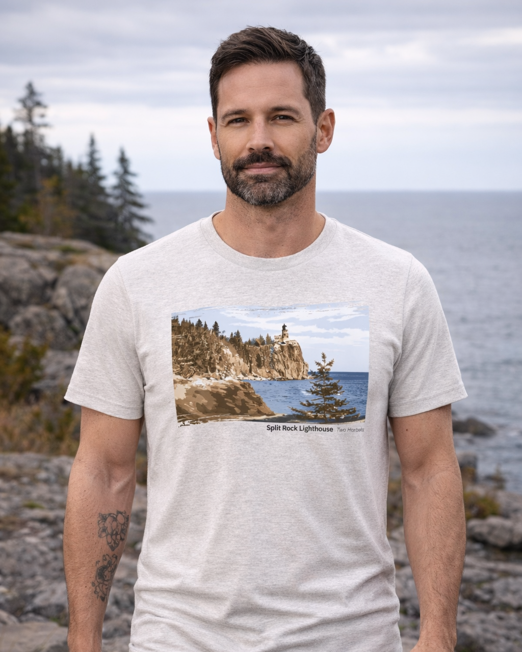 Man wearing a t-shirt with a scenic design of a lighthouse, standing outdoors by a body of water.